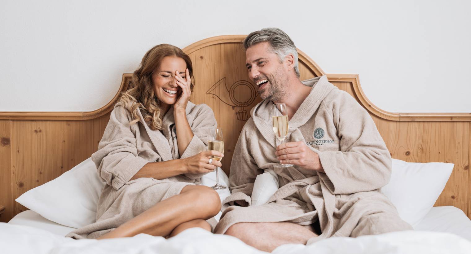 Paar sitzt auf einem Hotelbett und lachen bei einem Glas Sekt in der Hand