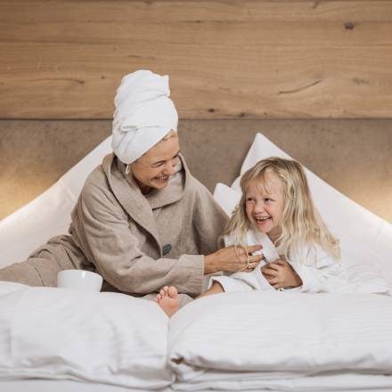 Mutter und Tochter sitzen mit Bademantel im Hotelbett und haben Spaß