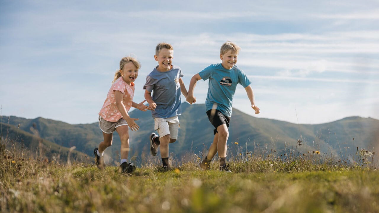 Drei Kinder rennen fröhlich über eine Wiese.