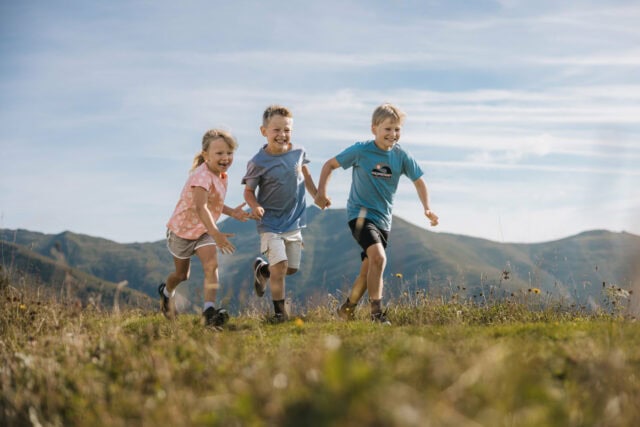 Drei Kinder rennen fröhlich über eine Wiese.